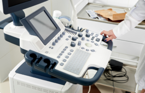 Ultrasound imaging machine with a control panel and a clinician's hand on the console/mode selector set in a clinical setting.