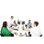 Five medical professionals sit around a circular table, using a central medical training device with screens and hand controls.