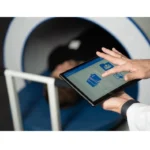 Healthcare professional uses a tablet with clinical icons beside an MRI scanner and patient, implying remote data access or control of the scan.