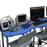 Medical monitoring station on a blue table with a patient monitor, ventilator, and computer displaying charts/graphs.