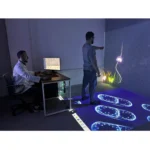 Two men in a lab: one stands and interacts with a wall projection of stars while glowing footprints appear on the floor; the other sits at a desk watching a computer screen.