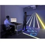 Two men in a dim room use a projection-based interactive display; one sits at a desk with a monitor while the other points at glowing lines projected on the wall and floor.
